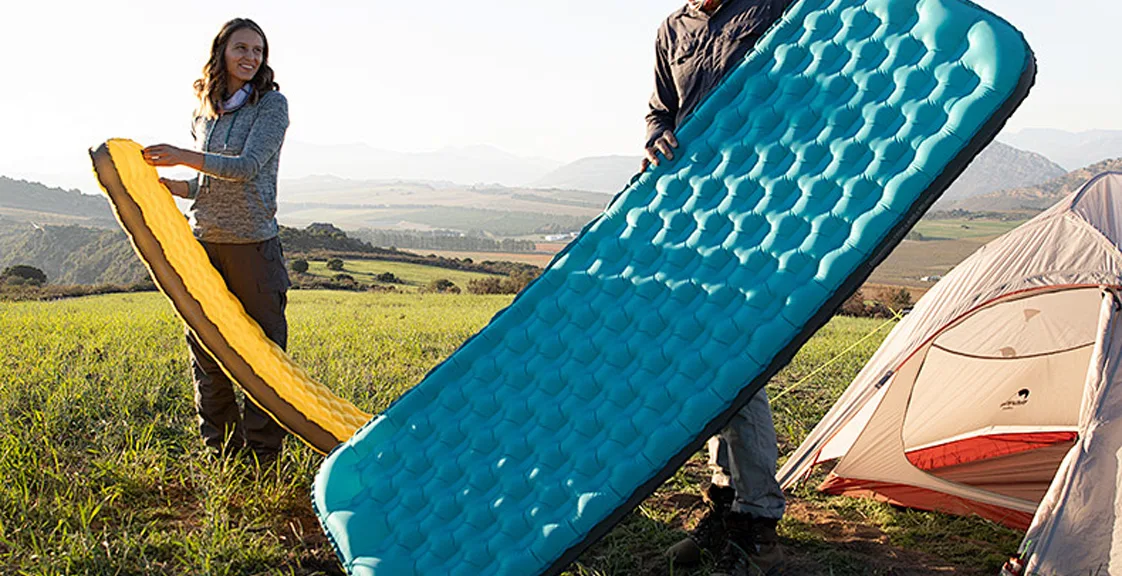 Campers holding inflatable camping air mattresses near a tent.